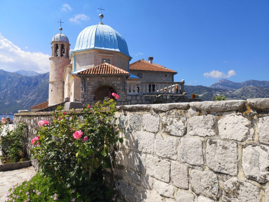 Landscape view on a sunny day of Our Lady of The Rocks Church with a blue roof and pink flowers in front at Our Lady of the Rocks Island in the Bay of Kotor, Montenegro