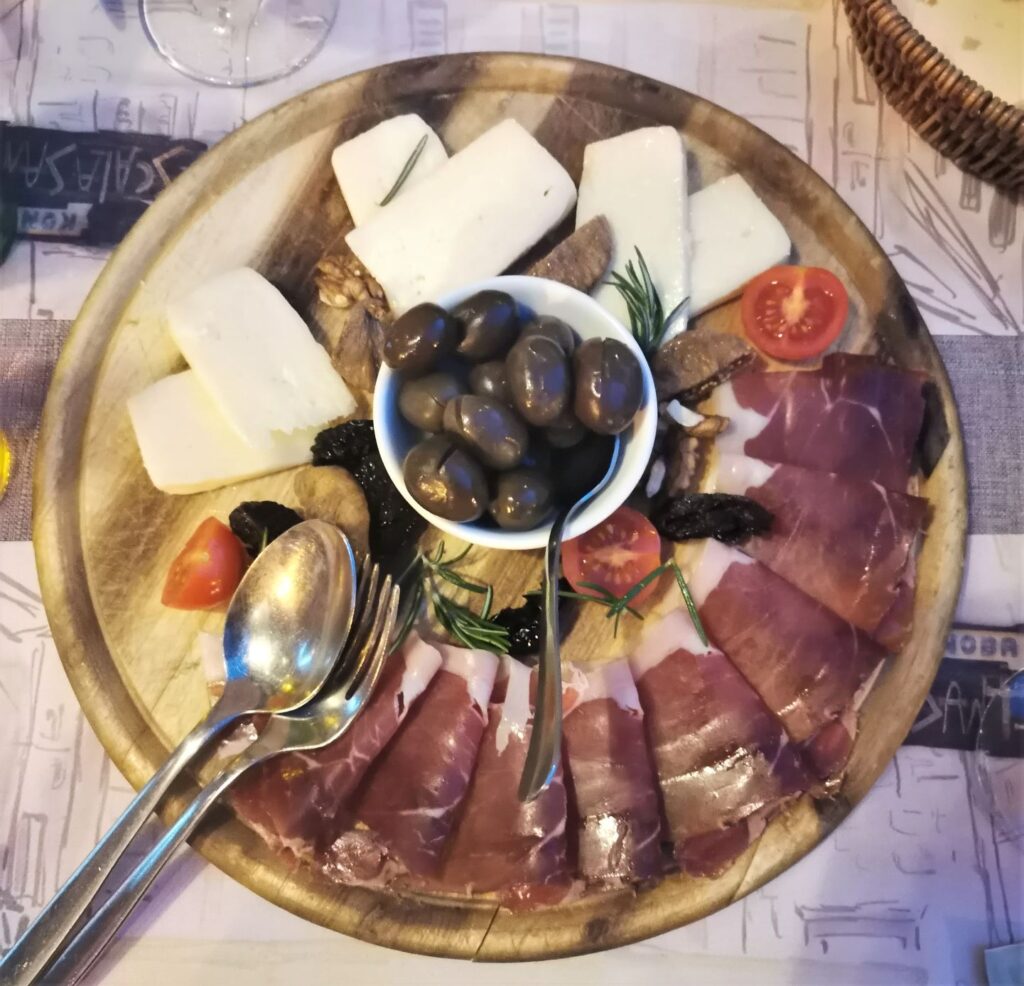 A large circular wooden board filled with Montenegro food and national dish of Njeguški Pršut and Montenegro cheese alongside olives, dried fruit and walnuts