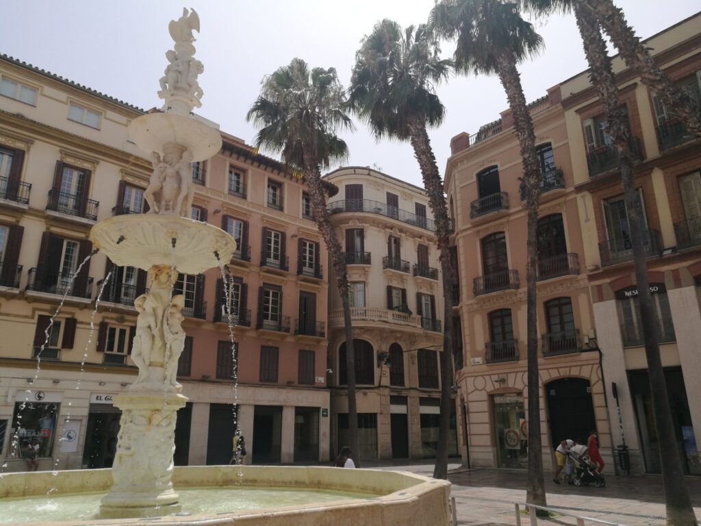 A decorative fountain stands in front of old buildings and towering palm trees in the old city streets of Malaga, Spain