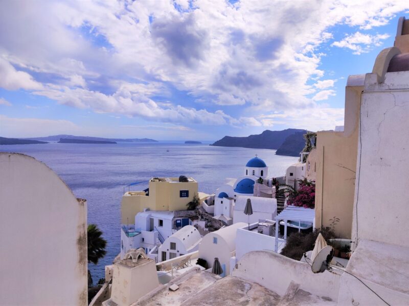 Scattered clouds in a blue sky over the town of Oia in Santorini, with white washed buildings on the edge of the cliffs. One of the best Greek islands to visit