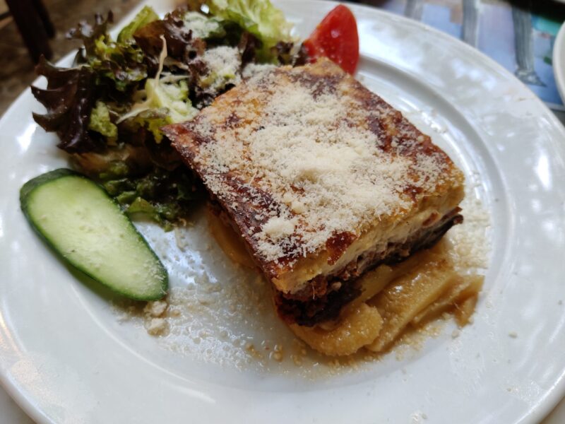 A white plate of moussaka with a small side salad and scattering of cheese