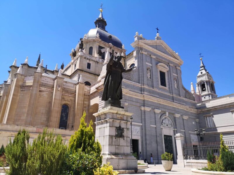 A beautiful blue sky day over Almudena cathedral in Madrid, Spain with statue and greenery in front of the cathedral entrance