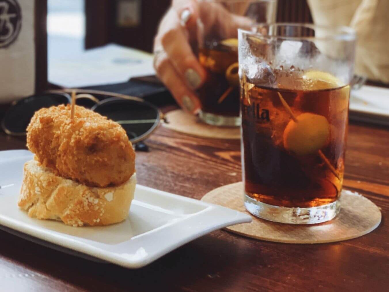 Two drinks with a plate of croquette on bread, like a pintxo